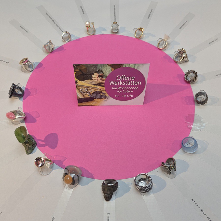 Rund um einen pinkfarbenen Papierkreis sind 20 einzelne Ringe auf weißem Hintergrund angeordnet. Es sind sehr verschiedene Ringe, jeder mit anderen Edelsteinen oder Materialien. Innen im pinken Kreis sieht man eine Werbe Karte der Offenen Werkstätten für das Wochenende vor Ostern, mit der Öffnungszeit der Werkstätten an diesem Wochenende, 10 - 19 Uhr.